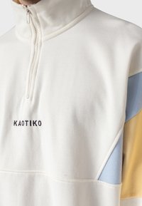 Sudadera de media cremallera en color crema con detalles en azul y amarillo. Tejido suave, acabado texturizado y logo bordado en el pecho.