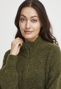 Maglione verde a maglia con colletto alto, dotato di una cerniera frontale e una finitura testurizzata. Gli accenti includono un anello argento per tirare la cerniera.