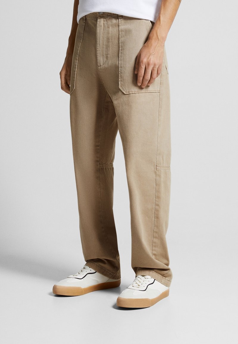 Homme portant un pantalon beige droit avec de grandes poches avant et des baskets blanches à semelles en gomme sur fond uni.