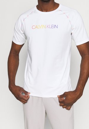 Mężczyzna w białej koszulce z krótkim rękawem z wielokolorowym logo "CALVIN KLEIN" na piersi, trzymający brzeg koszulki, widoczne szare spodnie.