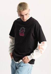 Camiseta de algodón negra con un logo gráfico que presenta un diseño puntiagudo y acentos en rosa, superpuesta sobre una camiseta de manga larga blanca con patrones en naranja.