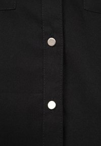 Camicia in tessuto nero con una fila verticale di cinque bottoni a scatto argentati. Texture liscia con dettagli di cucitura sottili.
