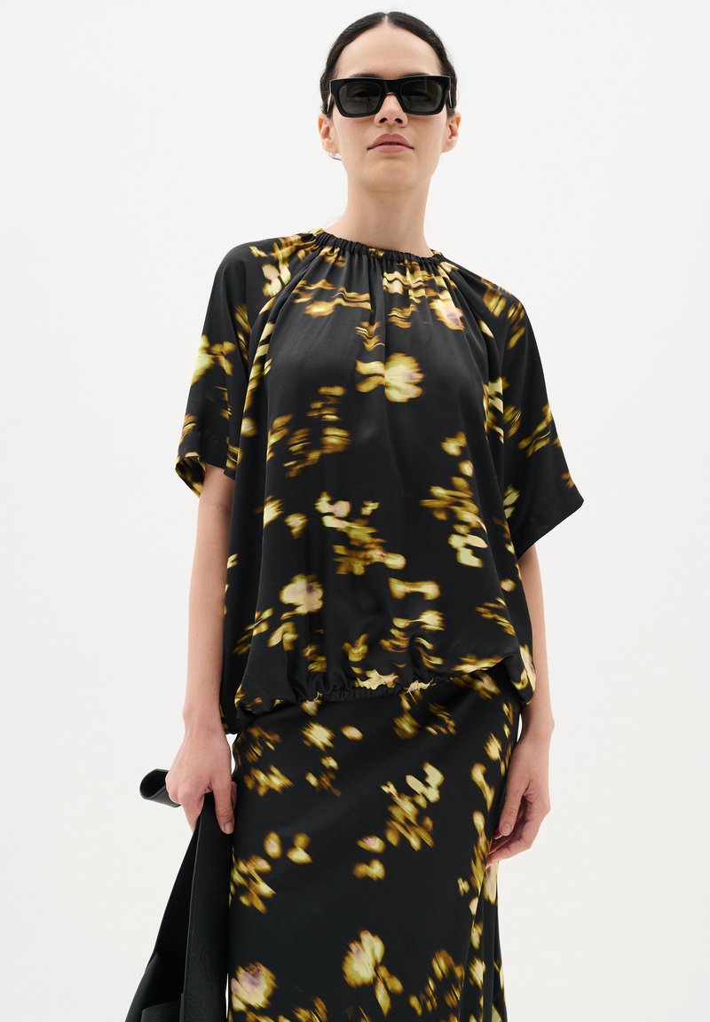 Blusa nera fluida con motivi floreali gialli, scollo elastico e maniche corte, abbinata a una gonna coordinata. Tessuto liscio e leggero.