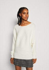 Pull en tricot blanc à côtes avec un large col et des manches longues, associé à une jupe texturée noir et blanc.