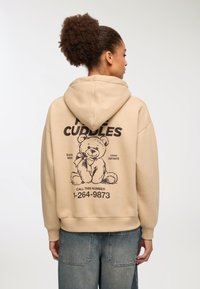 Beżowa bluza z kapturem z przewleczonym sznurkiem, na plecach z dużym czarnym nadrukiem misia pluszowego, towarzyszącym tekstem i numerem telefonu.