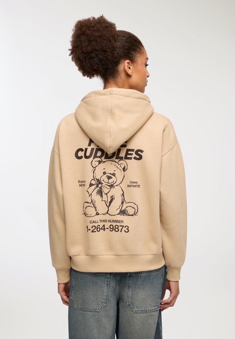 Beżowa bluza z kapturem z przewleczonym sznurkiem, na plecach z dużym czarnym nadrukiem misia pluszowego, towarzyszącym tekstem i numerem telefonu.