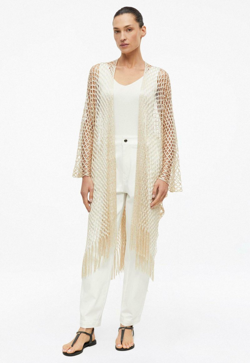 Woman wearing white top and pants with beige open-knit shawl featuring fringe and black T-strap sandals, standing against plain background.