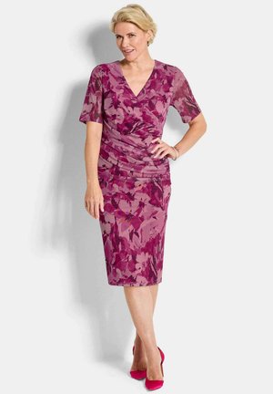 Blonde Frau steht mit einer Hand in der Hüfte, träg ein knielanges, kurzärmeliges Kleid mit rosa- und lila Muster sowie passende High Heels.