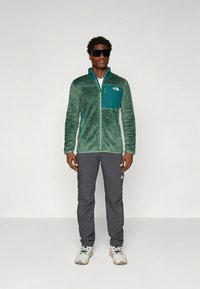 Groene fleece jas met een teal borstzak, gecombineerd met grijze broek en witte schoenen. Voorzien van een ritssluiting en een gestructureerde stof.