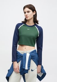 Sötétzöld crop top hosszú tengerészkék ujjal, fehér díszítéssel és egy kis logóval. Bézs nadrággal és a derekán megkötött kék kabáttal párosítva.