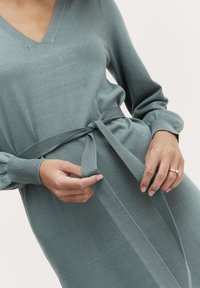 Abito in maglia di un teal smorzato, con scollatura a V, maniche lunghe e una cintura in tessuto legata in vita. Materiale testurizzato e silhouette aderente.