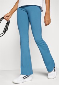 Kék, bővülő leggings rugalmas anyagból, sima felülettel és oldalsó varrással, fehér sportcipővel párosítva.