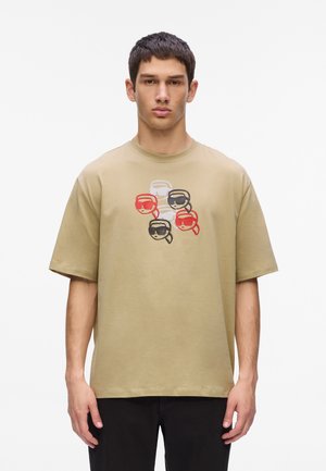 Camiseta de algodón beige con un corte relajado. En el frente presenta un gráfico de cuatro caras estilizadas con gafas de sol de colores rojo, negro, blanco y gris.