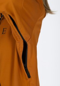 Giacca arancione con tasca con zip sul lato sinistro, realizzata in tessuto liscio con dettagli di cucitura visibili. Zip nera e dettagli in nero.