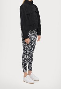 Veste noire courte avec deux poches avant, associée à des leggings blancs ornés d'un imprimé léopard noir, et des baskets blanches avec des semelles beiges.