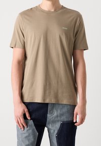 Mand iført en beige HUGO T-shirt og patchwork jeans i mørkeblå og lyseblå mod en ensfarvet baggrund.