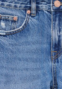 Jeans de denim azul claro con una textura desgastada, que presentan herrajes de cobre, bolsillos cosidos y deshilachados visibles en el borde del bolsillo.
