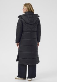 Melnā pūkainā jaka ar kapuci, ar garu, quilted dizainu, elastīgām piedurknēm un sašaurināta vidukļa detaļu.