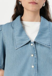 Lichtblauwe denim shirt met een grote kraag, korte mouwen en knopen aan de voorkant. Gelaagd stof met subtiele stikseldetails.