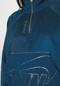 Blå zip-up tröja med texturerad tyg. Har guld "Just do it"-text och en reflekterande Nike-logotyp på framfickan.