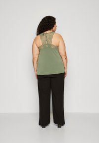 Olivengrünes Tanktop mit Spitzenrückendetail, verstellbaren Trägern, kombiniert mit lockeren schwarzen Hosen, die eine entspannte Passform und weiche Textur zeigen.