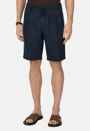 Homme portant un short bleu marine à cordon de serrage arrivant aux genoux et des sandales marron à brides croisées, debout devant un fond uni.