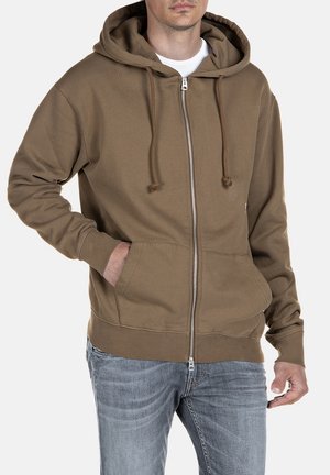 Mand iført brun hættetrøje med lynlås og snore samt blå jeans, med den ene hånd i lommen, stående mod en hvid baggrund.
