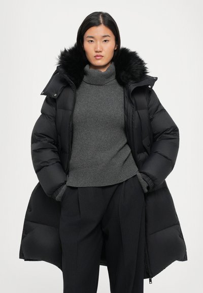 Schwarze Daunenjacke mit einer Kapuze aus Kunstfell, ausgestattet mit einem Reißverschluss und gesteppter Optik, kombiniert mit einem grauen Rollkragenpullover und einer maßgeschneiderten Hose.