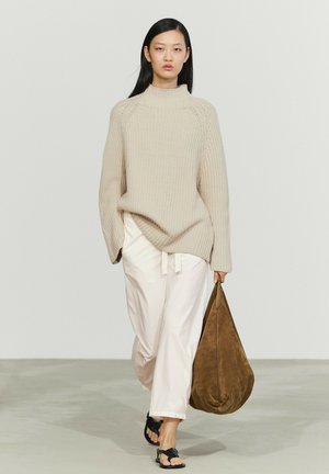 Donna con maglione beige a maglia, pantaloni bianchi larghi, sandali neri, che porta una grande borsa in suede marrone su uno sfondo semplice.