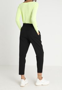 Mustat kapenevat housut, joissa on sivutaskut, yhdistettyinä pitkähihaiseen neonvihreään puseroon. Valkoiset tennarit täydentävät asua. Sileä kankaan rakenne.