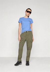 Γυναίκα στέκεται με τα χέρια χαλαρά, φορώντας μπλε μπλουζάκι, ελιά πράσινα παντελόνια cargo, μαύρες ορειβατικές μπότες και μεγάλα μαύρα γυαλιά ηλίου σε λευκό φόντο.