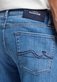 Jasnoniebieskie dżinsy z tylną kieszenią z ciemnoniebieskim wzorem w zygzak. Zawierają granatową metkę z napisem "7 For All Mankind".