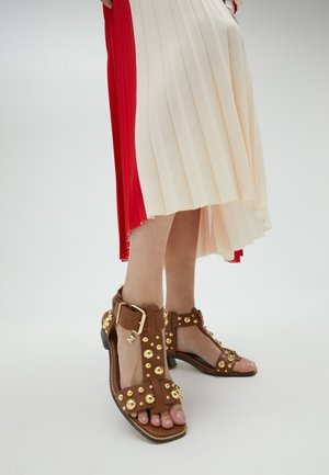 Sandales brunes à brides avec des clous ronds dorés, bride de cheville réglable et petit talon. Les pieds sont positionnés devant une robe plissée.