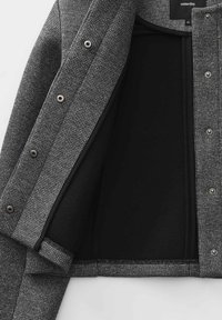 Giacca grigia con texture e fodera interna nera, caratterizzata da chiusure con bottoni a pressione e un design aderente. Gli accenti includono bordi in contrasto.