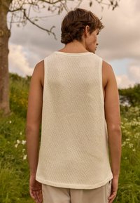 Lys beige ærmeløs tanktop med et struktureret, åndbart strikkemønster. Kantene er let buede og har en kontrasterende hvid halsudskæring og ærmeåbninger.