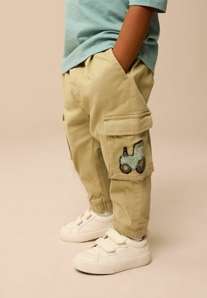 Beige Cargo-Hosen mit grüner Traktor-Stickerei am rechten Bein, kombiniert mit weißen Sneakers und einem hellblauen Hemd.