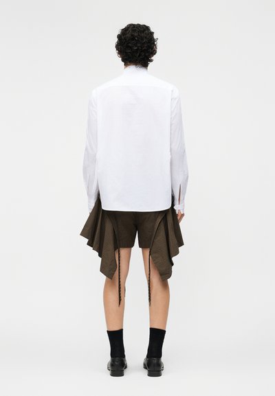 Osoba o kręconych włosach widziana od tyłu, ubrana w białą koszulę z długim rękawem i brązowe asymetryczne spodenki, czarne skarpetki i buty.