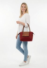 Heledate juustega naine valges pluusis ja teksades hoiab kahest sangast suurt punast ja pruuni käekotti, naeratab ning seisab valgel taustal.