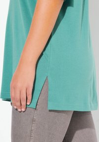 Kurzärmliges türkisfarbenes Shirt mit relaxed fit, seitlichen Schlitzen und flachen Nähten, kombiniert mit grauen Hosen. Weiches Baumwollmaterial.