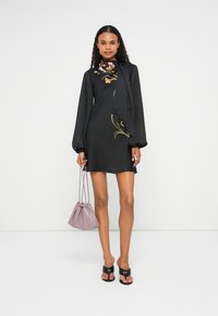 Rochie neagră cu mâneci lungi tip puff, având accente florale în nuanțe de aur și roz. Modelul poartă o geantă lavandă și sandale negre.