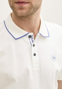 Polo bianca con colletto bordato di blu, pattina con tre bottoni, bottoni blu scuro e un piccolo logo sul lato sinistro del petto. Tessuto texturizzato.