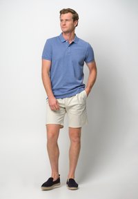 Blå pikétröja, beige shorts och mörka slip-on skor. Skjortan har krage och korta ärmar; shorts är skräddarsydda och lätta.