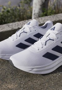 White running shoes with navy blue Adidas stripes placed outdoors on a textured surface near a low concrete wall.