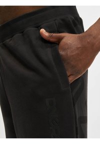 Svart joggebukser med ribbestrikket midje og sidelommer. En matt svart logo vises på venstre ben, med en jevn tekstur.