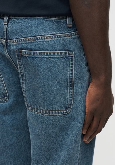 Δεξί χέρι ακουμπισμένο δίπλα σε ανοιχτόχρωμο τζιν παντελόνι με μεγάλη πίσω τσέπη, ορατές θηλιές για ζώνη και μαύρο πουκάμισο περασμένο μέσα.