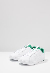 Baskets en cuir blanc avec doublure intérieure verte et languettes, logo crocodile perforé sur le côté, posées sur une surface blanche.
