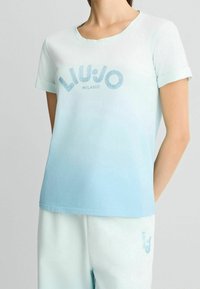 Camiseta de algodón azul claro con mangas enrolladas, que presenta un diseño de texto en blanco y azul "LIU JO MILANO", desvaneciéndose a un azul más oscuro en el dobladillo.