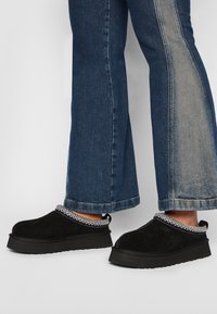 Black suede slip-on shoes with a textured knit collar and a thick black sole, paired with wide-leg blue jeans featuring a contrasting panel.