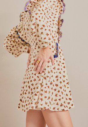 Femme portant une robe fleurie crème avec de longues manches bouffantes et un bord festonné bleu, les mains posées sur les hanches, bagues en or aux doigts.