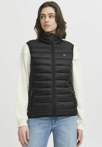 Gilet en doudoune noir avec col haut, motif matelassé horizontal, fermeture éclair sur le devant et un petit logo sur la poitrine. Porté sur un haut de couleur claire.